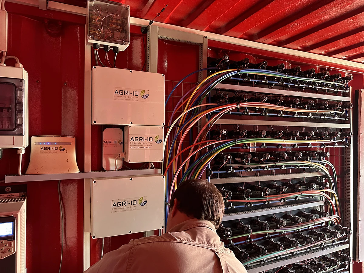 The image shows a man working inside a control unit of an automated farming system. The interior is filled with a series of electrical components, wiring, and devices labeled AGRI-IO which appear to be part of a smart automation system for agricultural management. The wall is painted red, and several cables of various colors are connected to a large array of valves or similar devices, indicating a sophisticated setup for controlling irrigation or other automated farm functions. The man is facing away from the camera, focusing on his work.
