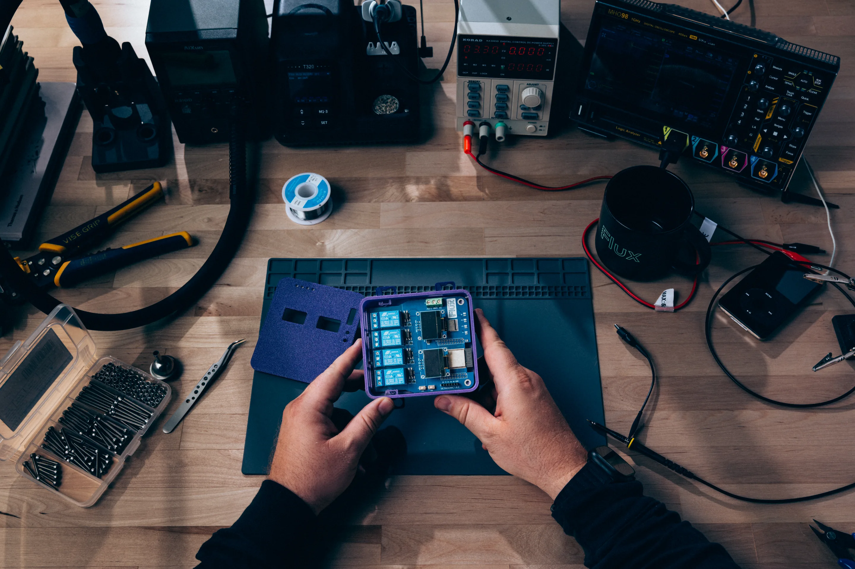 Hands holding a blue relay circuit board over an electronics workbench