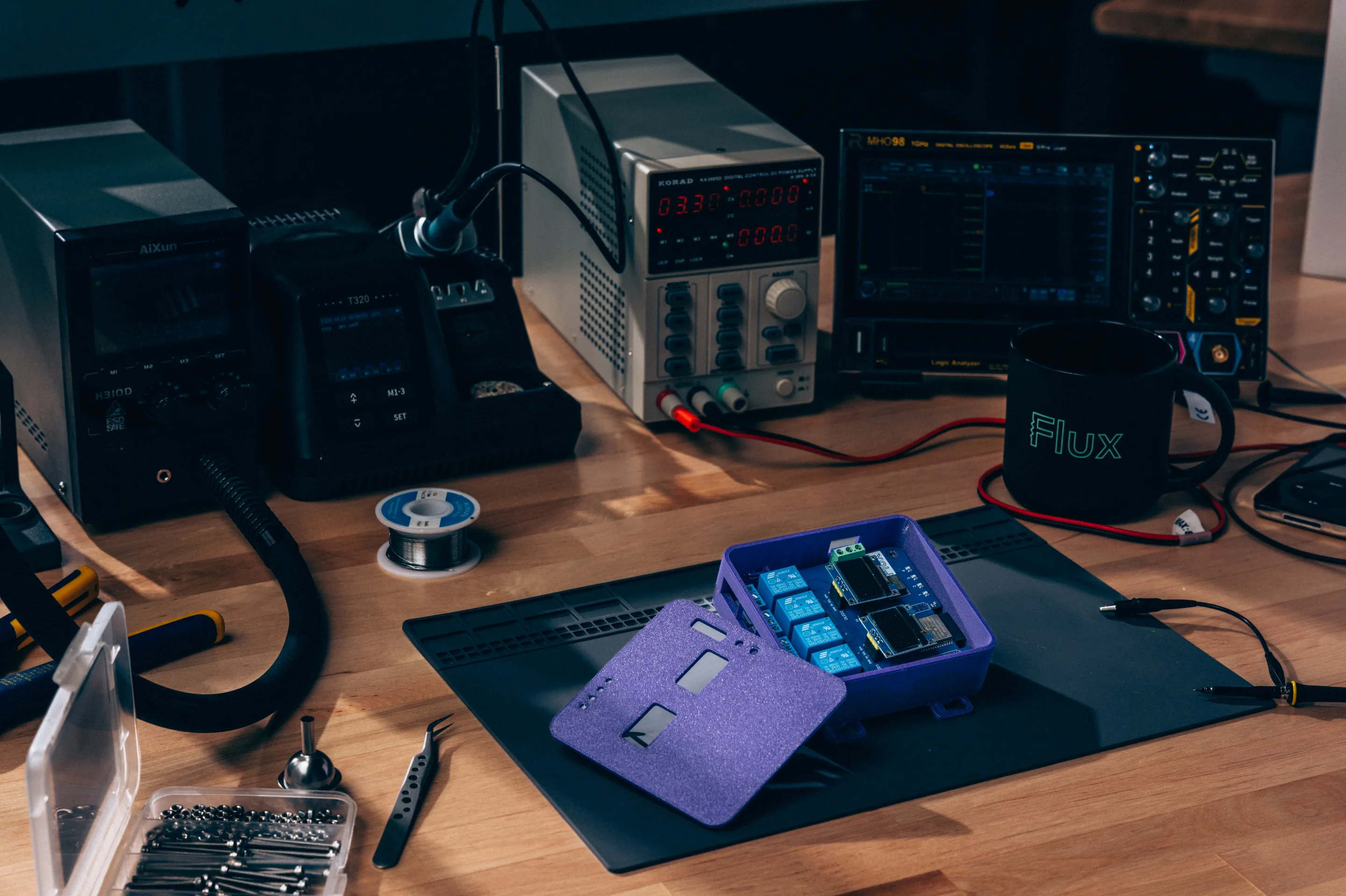 A purple enclosure with a relay board on an ESD mat, with bench power supplies and an oscilloscope behind it